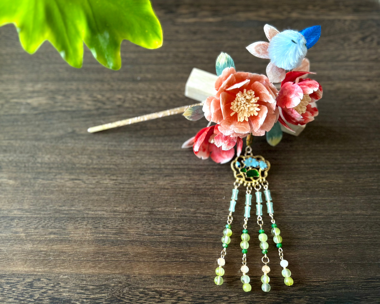 Detail shot of a meticulously crafted hair accessory featuring velvet flowers, aka Rong hua/Ronghua Traditional Chinese design, and a delicate beaded dangle. presented on a dark wooden backdrop.