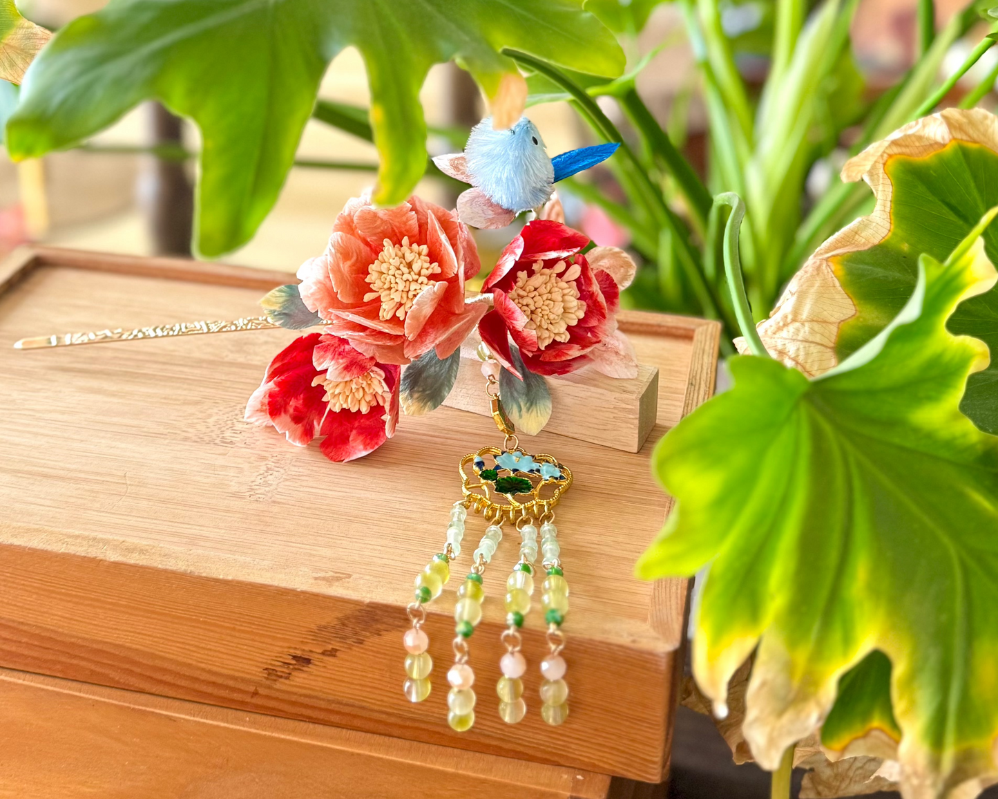 Detail shot of a meticulously crafted hair accessory featuring velvet flowers, aka Rong hua/Ronghua Traditional Chinese design, and a delicate beaded dangle. presented on a yellow wooden backdrop with a green plant behind it.
