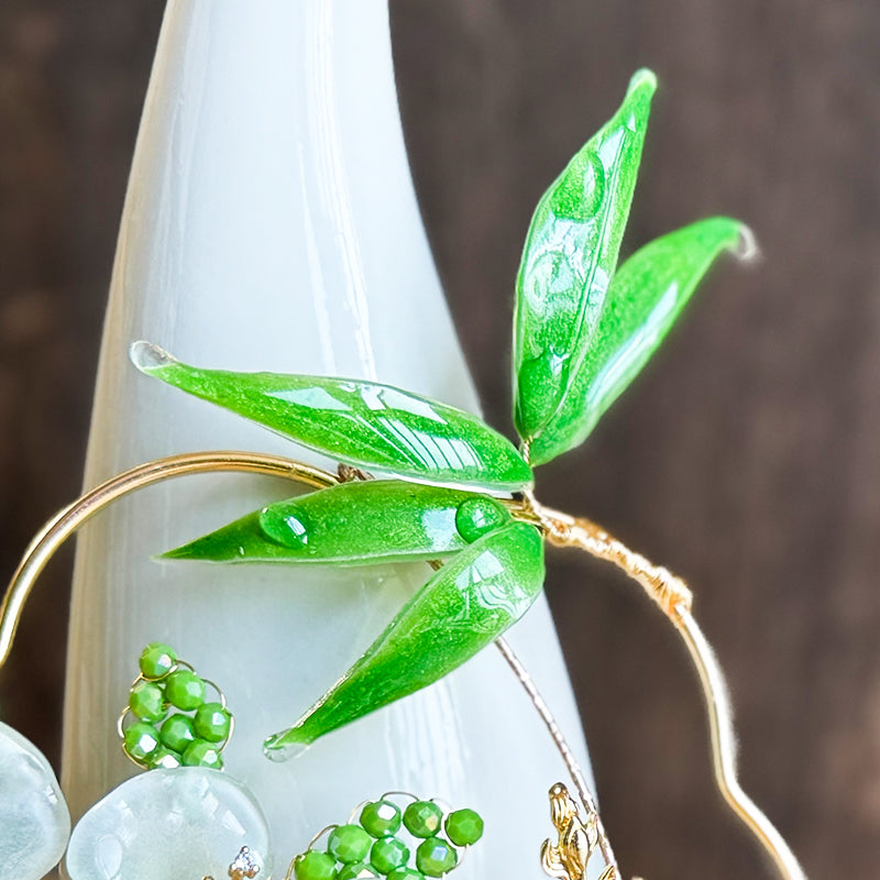 The image shows a close-up of a piece of craft work, which is the skeleton of a handmade fan, featuring delicate green glass or crystal leaves.