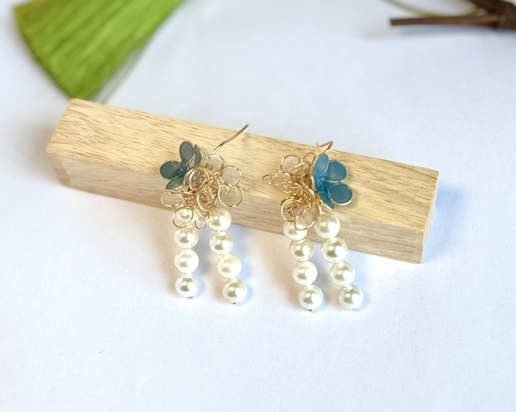 Elegant handcrafted earrings featuring blue sea glass flowers with gold wire accents and dangling pearl strands, displayed on a light wooden block against a white background with a subtle green accent visible.