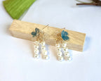 Elegant handcrafted earrings featuring blue sea glass flowers with gold wire accents and dangling pearl strands, displayed on a light wooden block against a white background with a subtle green accent visible.
