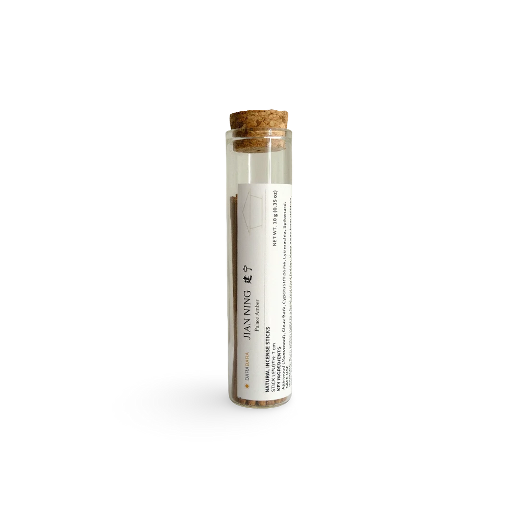 A clear glass tube with a cork stopper and a white label reading JIAN NING Incense, containing brown herbal powder, photographed on a clean white background.