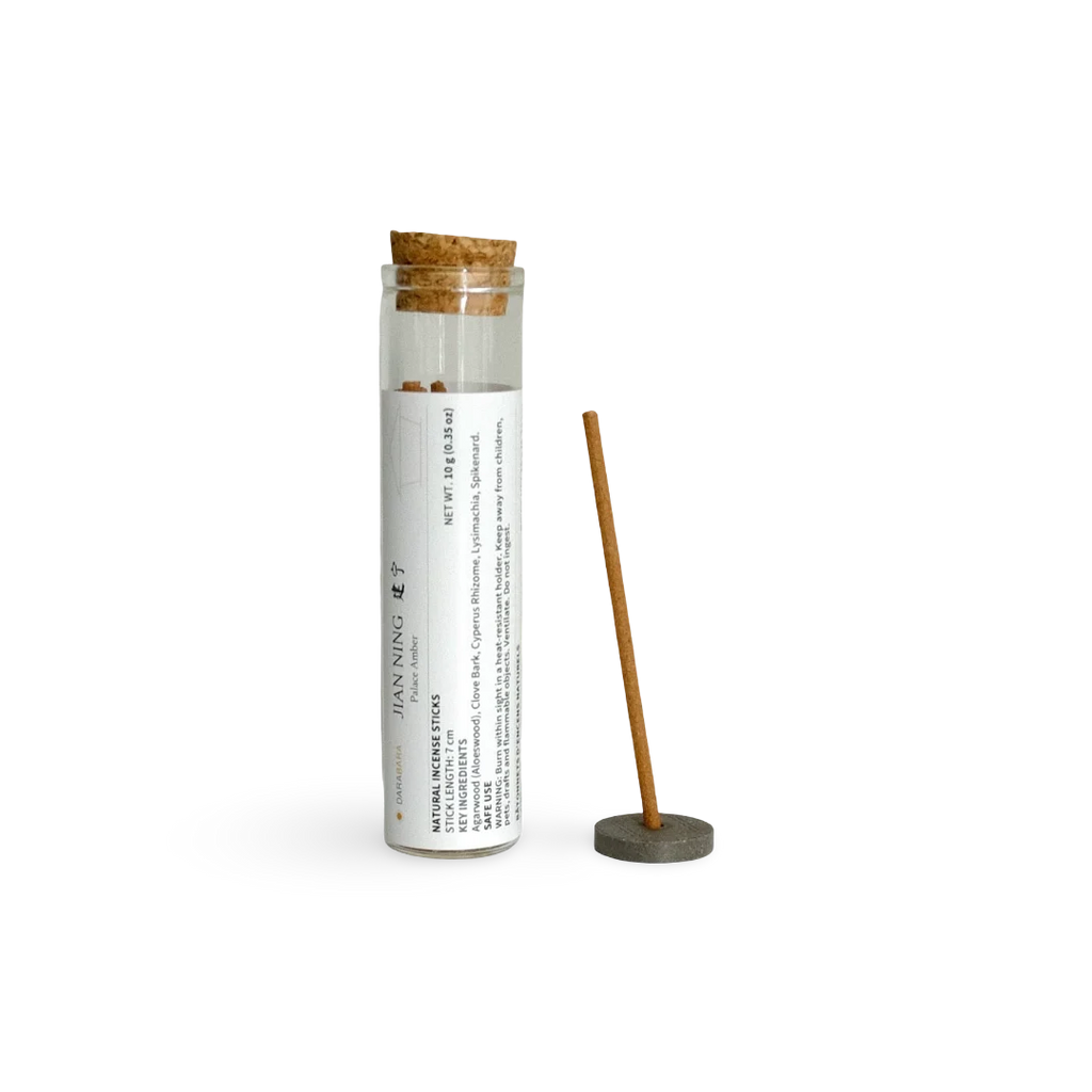 A bamboo incense stick displayed next to its clear glass storage tube with a cork stopper and a white printed label. The tube is labeled 'JAN NING' and 'NATURAL INCENSE STICK'.