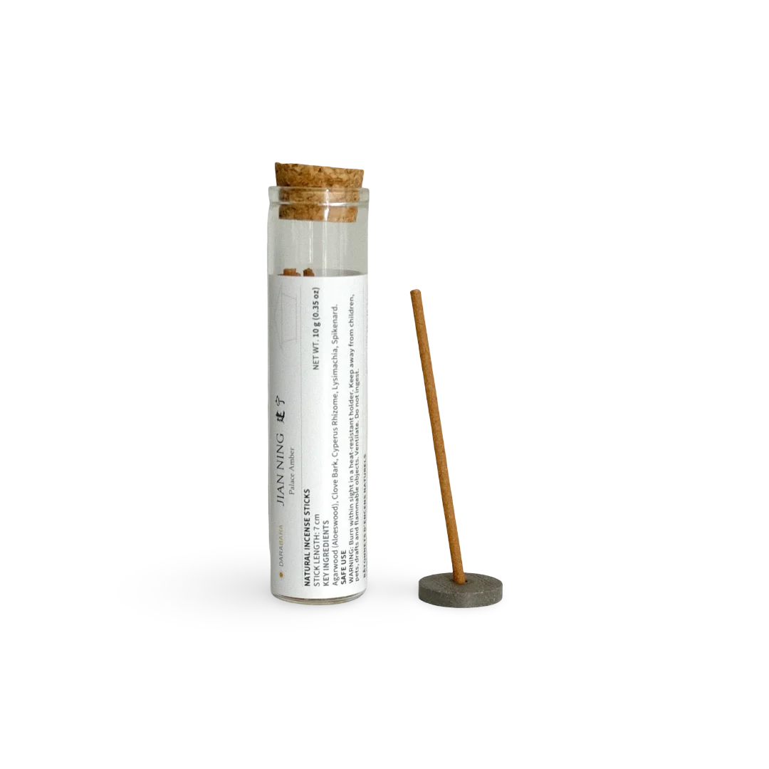 A bamboo incense stick displayed next to its clear glass storage tube with a cork stopper and a white printed label. The tube is labeled 'JAN NING' and 'NATURAL INCENSE STICK'.