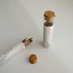 Two clear glass vials with cork stoppers containing brown incense sticks of Palace Amber by Darabara, shown on a light gray surface. One vial is upright and the other lies on its side, with a separate cork stopper placed in front.