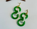 Duplex Viridis luxury drop earrings featuring double emerald green jade hoops connected by elegant freshwater pearl strands. Each earring showcases two genuine jade rings in vibrant forest green with a lustrous finish, artfully linked by white freshwater pearls in a curved design. Gold-plated posts and findings complement the modern Asian-inspired style. These statement earrings combine traditional jade with contemporary geometric design, perfect for both special occasions and elevated everyday wear. Handcr