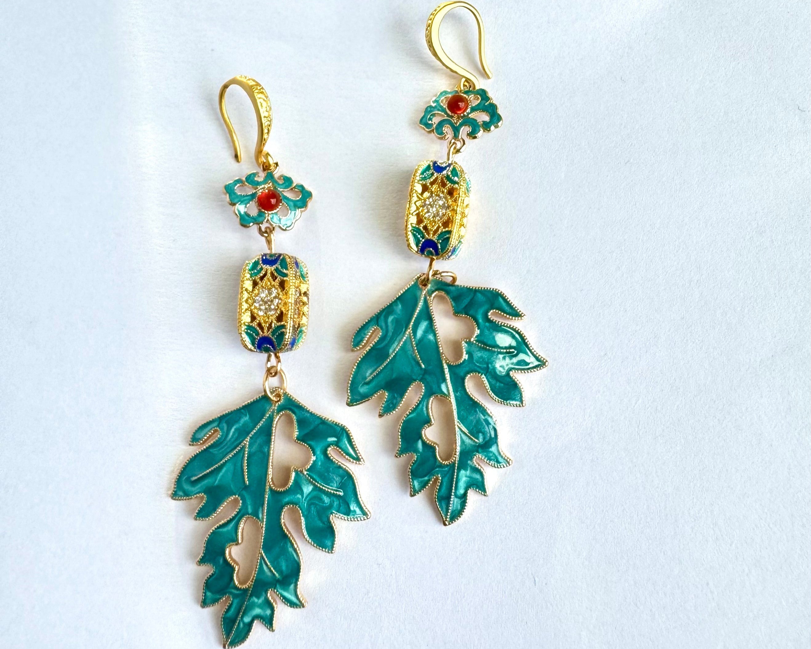 Elegant drop earrings featuring teal enamel oak leaves with gold trim, connected to decorative rectangular charms with blue accents and topped with ornate teal and red floral hooks.