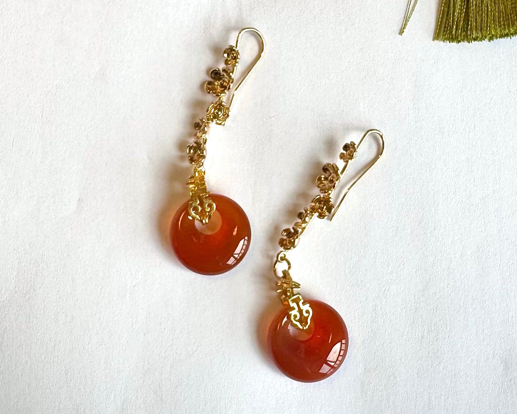 Close-up overhead shot of two elegant, handmade "Ruby Moon" earrings on a white surface. The earrings feature golden alloy frames with delicate petal-like details, dangling red agate rings that resemble solid rubies, and small red bead accents. A green tassel is partially visible in the upper left corner.