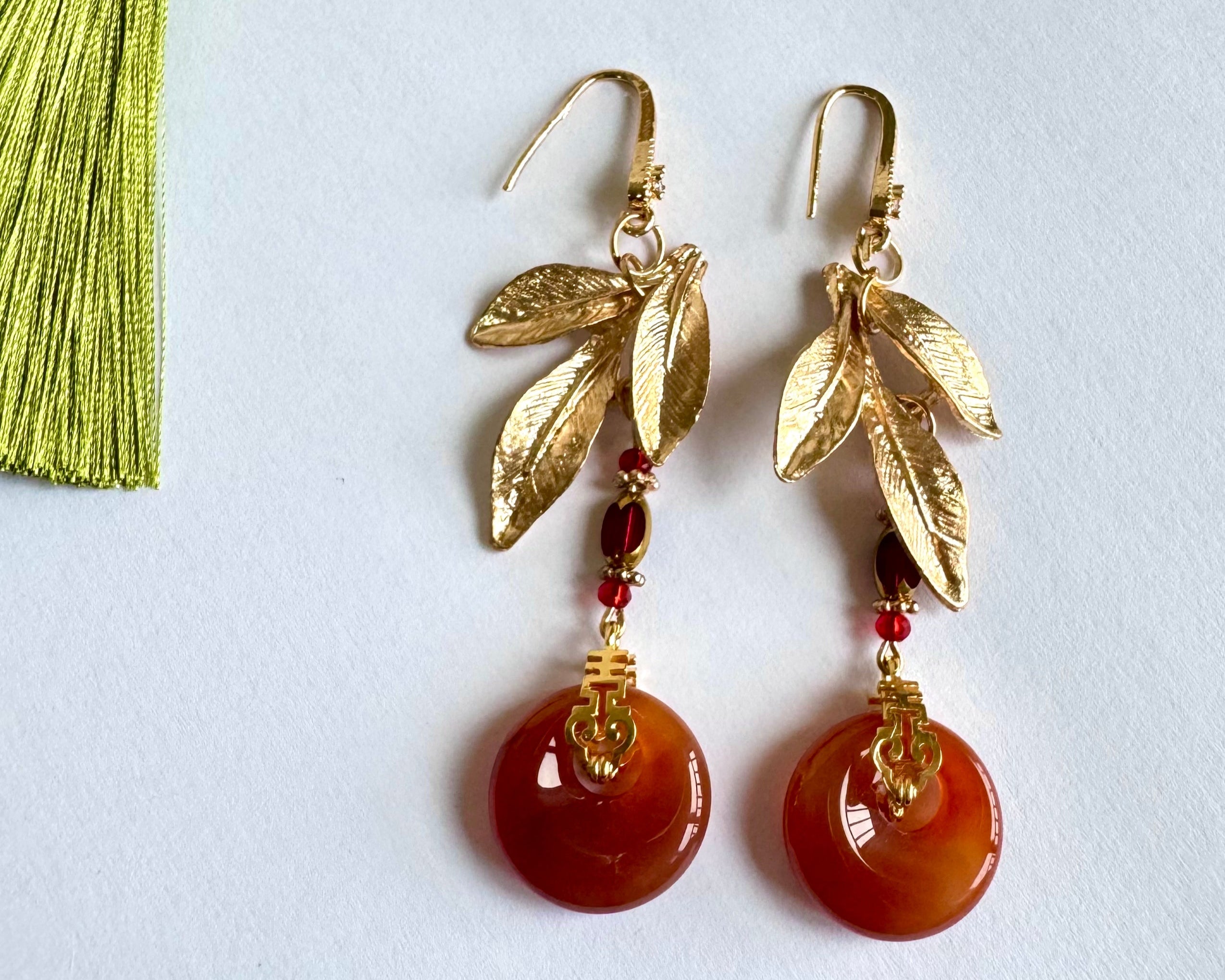A detailed view of handcrafted "Ruby Moon" earrings showcasing their golden alloy frames, polished red agate "ruby" rings, and subtle red bead embellishments. A soft green tassel lies nearby, emphasizing the artisanal nature of the jewelry on a clean white backdrop.