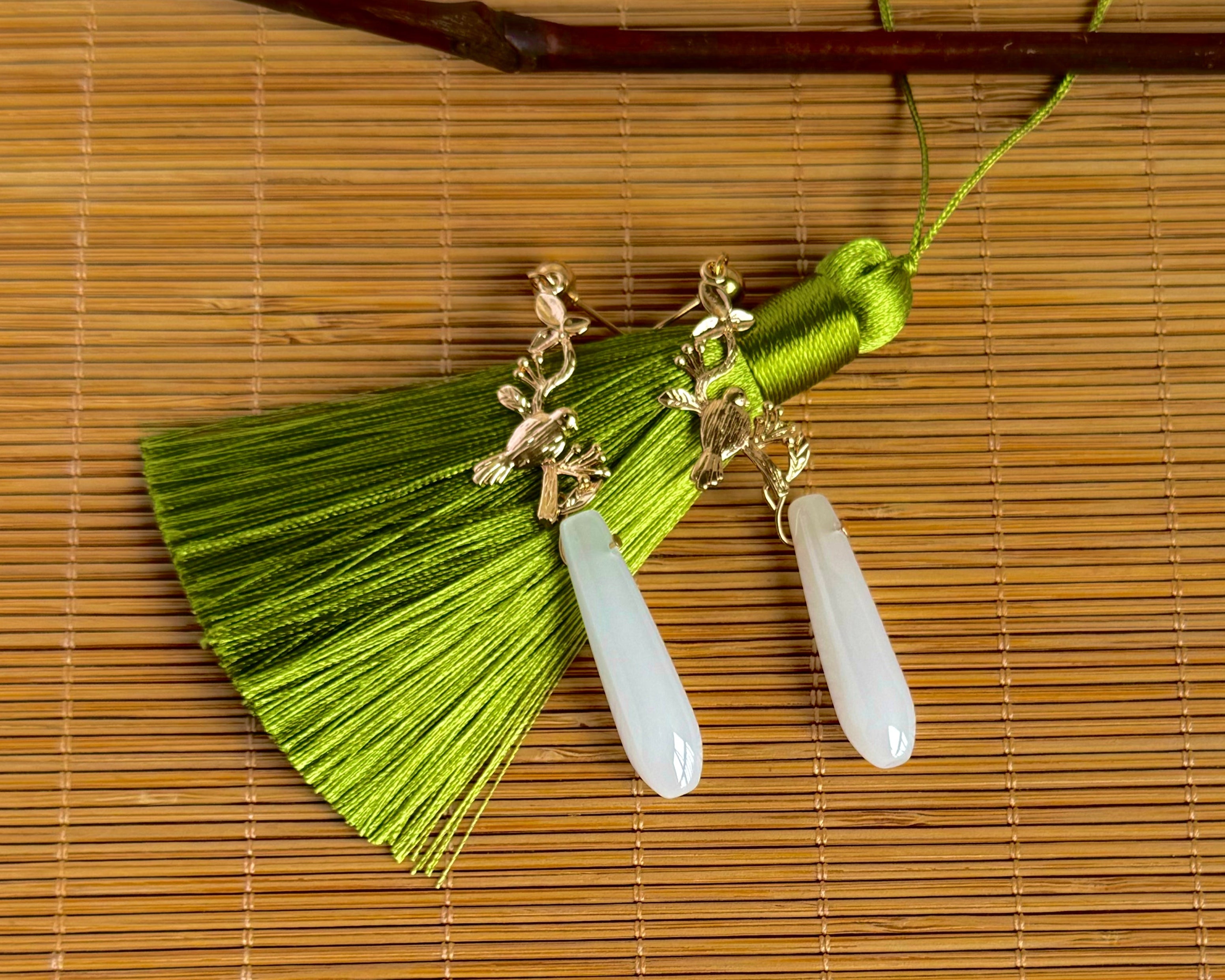 "Spring Silk statement tassel earrings featuring vibrant lime green silk tassels paired with elegant white jade drops. Gold-tone bird charm connectors add a whimsical touch. These lightweight boho-chic earrings combine natural stone and luxurious silk threads, perfect for spring and summer fashion. Each tassel measures approximately 2 inches in length, creating a dramatic yet wearable statement piece. Handcrafted design ideal for casual to dressy occasions."
