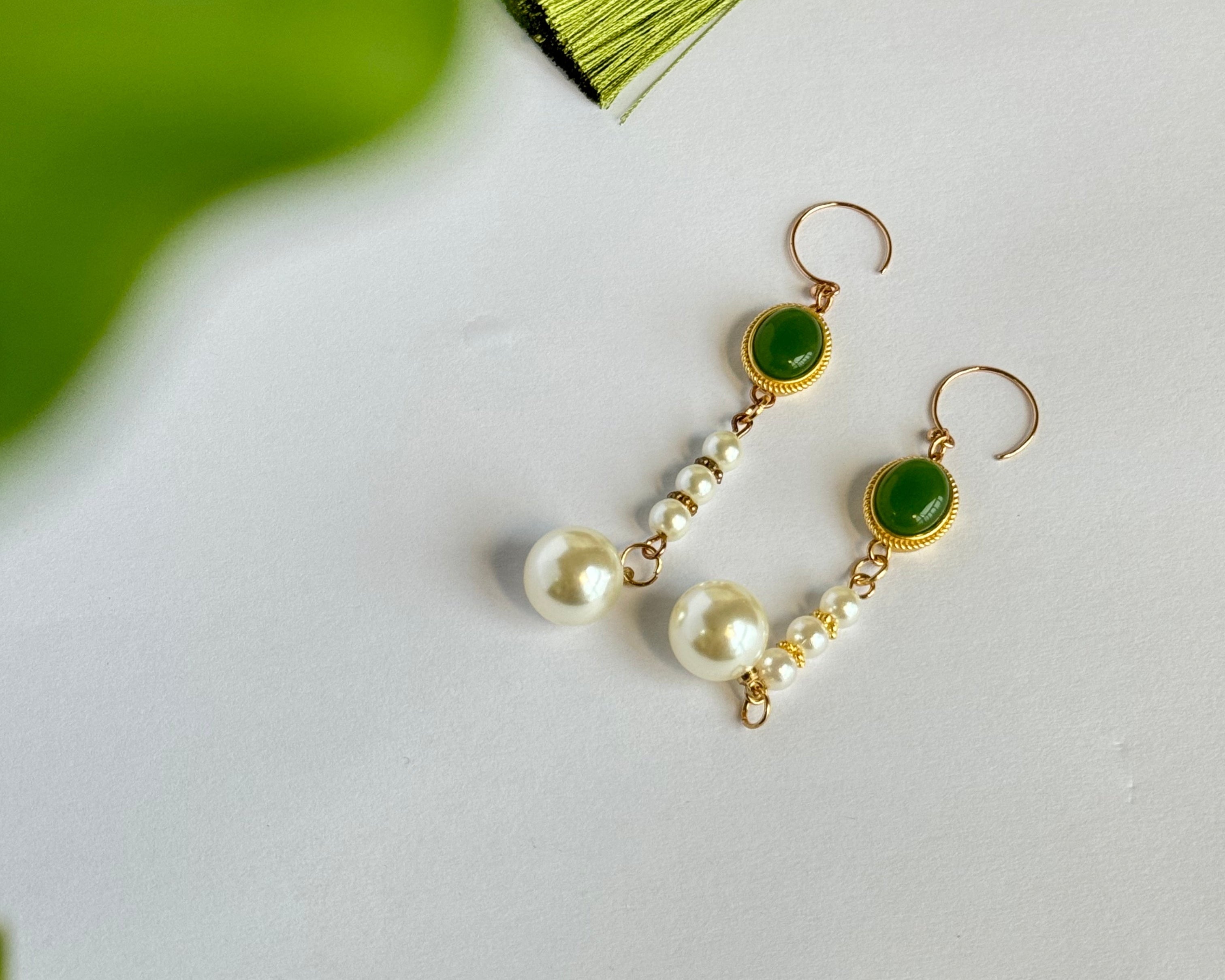Gold drop earrings featuring jade green stone settings with pearl accents - smaller pearls along the chain and larger pearl pendants at the bottom, displayed on a white surface with subtle green backdrop.