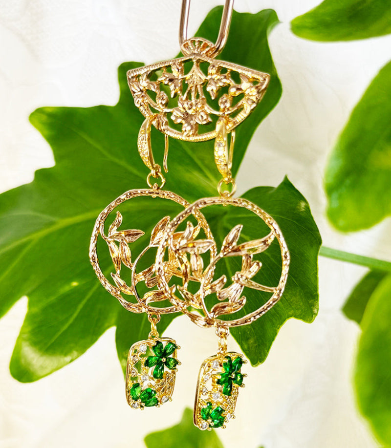 This image shows a beautiful pair of handmade earrings alongside a lime green tassel. The earrings feature gold-toned circular pendants with intricate leaf or branch designs, from which hang rectangular charms adorned with green gemstone elements that appear to be zircon.