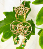 This image shows a beautiful pair of handmade earrings alongside a lime green tassel. The earrings feature gold-toned circular pendants with intricate leaf or branch designs, from which hang rectangular charms adorned with green gemstone elements that appear to be zircon.