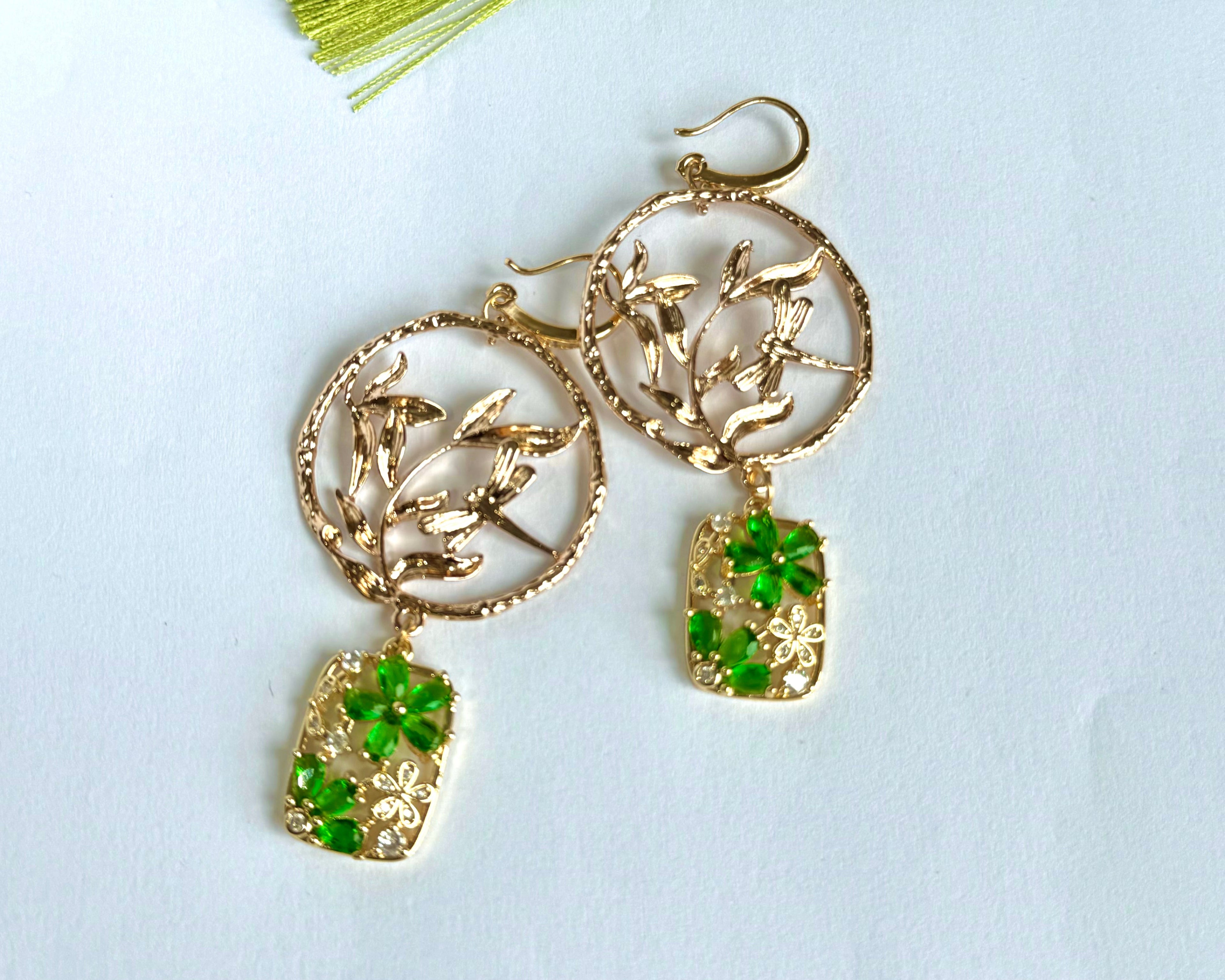 This image shows a beautiful pair of handmade earrings alongside a lime green tassel. The earrings feature gold-toned circular pendants with intricate leaf or branch designs, from which hang rectangular charms adorned with green gemstone element