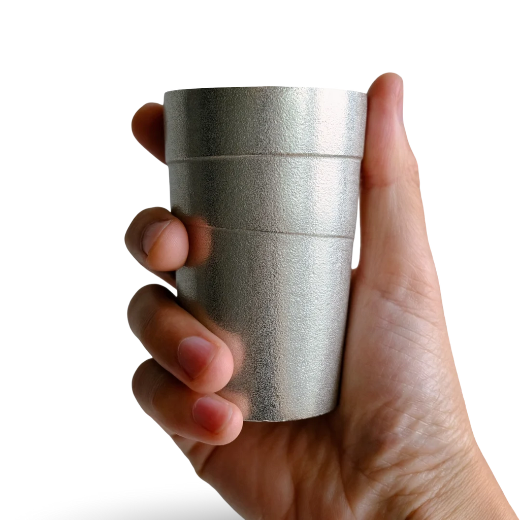 Hand holding a silver tin cylindrical beer tumbler with circular lines against a transparent background
