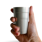 Hand holding a silver tin cylindrical beer tumbler with circular lines against a transparent background