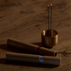 two kunjudo incense tubes laying on a wooden surface, along with a brass metal incense holder with two incense sticks both burning on the side