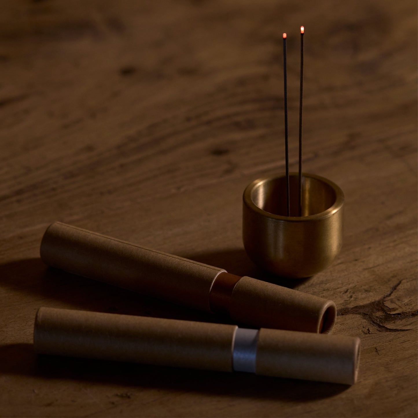 two kunjudo incense tubes laying on a wooden surface, along with a brass metal incense holder with two incense sticks both burning on the side