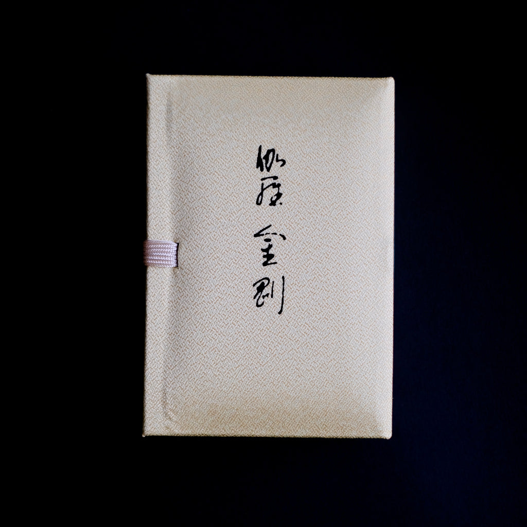 Nippon Kodo Kyara Kongo incense in its elegant, fabric-covered Paulownia wood box. The box is cream-colored with Japanese calligraphy, and a small purple ribbon is visible on the side. This premium Japanese incense is made from selected aloeswood and includes 60 sticks with a ceramic holder.